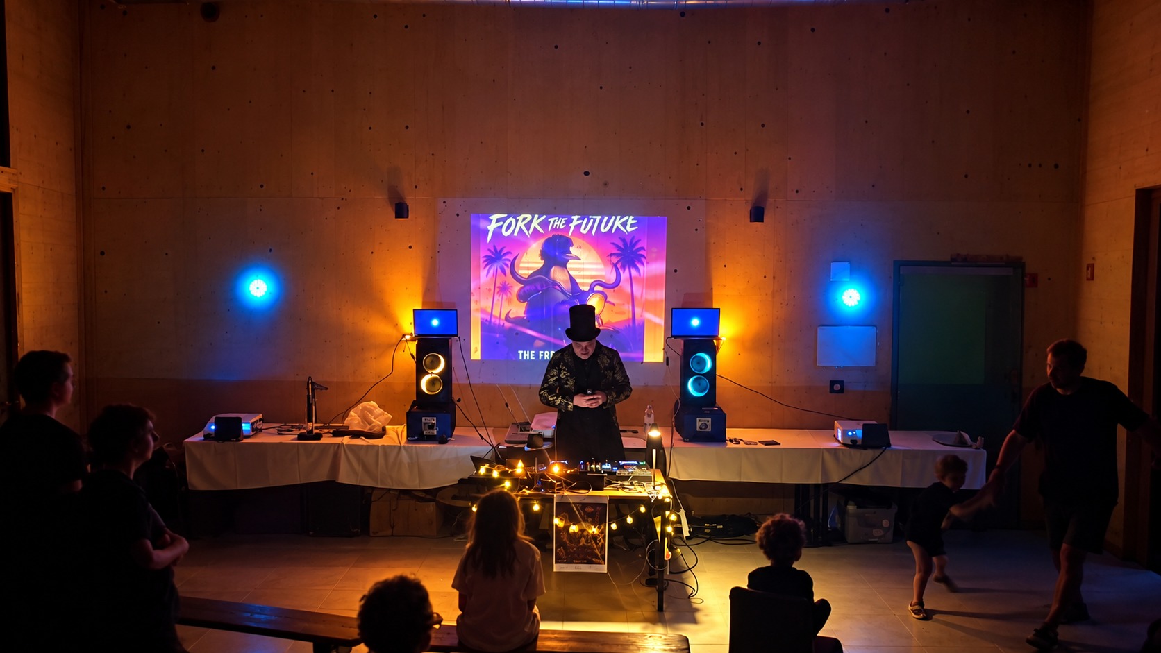 Jasper Nuyens in a top hat at the DJ table at Fri3d Camp, with the Fork the Future artwork projected on the wall behind him, laptops on two speaker towers, short-throw projectors on the side tables, lantern lights along the table and the audience in silhouette.