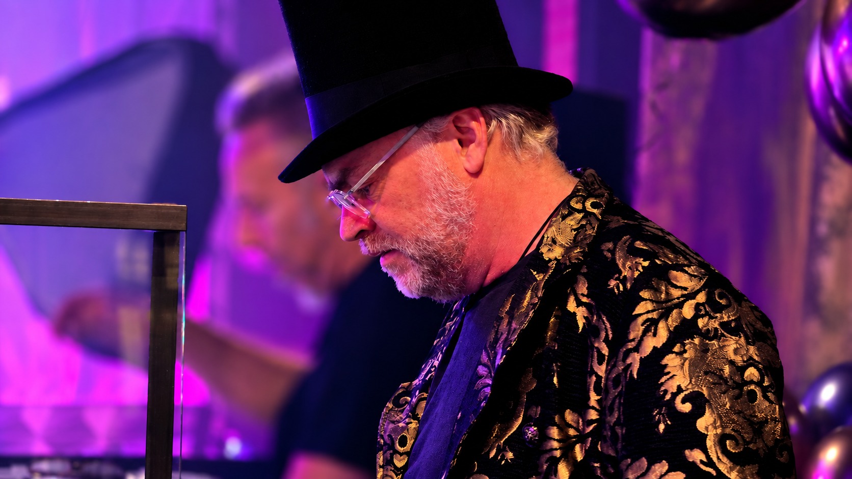 Jasper Nuyens in a top hat, round glasses and a black and gold brocade jacket, looking down at the decks under purple stage light, with a second person blurred in the background.